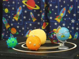 Zdjęcie przedstawia układ słoneczny zrobiony przez dzieci. Znajduje się w nim pomarańcza, nimi globus i różne zabawki.
