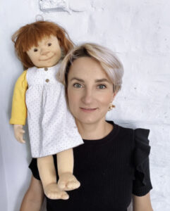 Zdjęcie młodej kobiety w krótkich blond włosach, trzymającą na ramieniu własnoręcznie wykonaną lalkę.