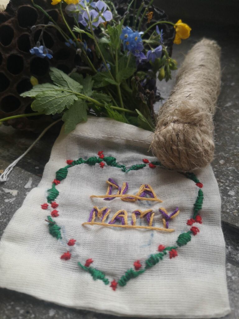 Zdjęcie przedstawia beżowy woreczek z wyhaftowanym sercem i napisem Dla Mamy. Obok leży polny bukiet kwiatów oraz motek sznurka.