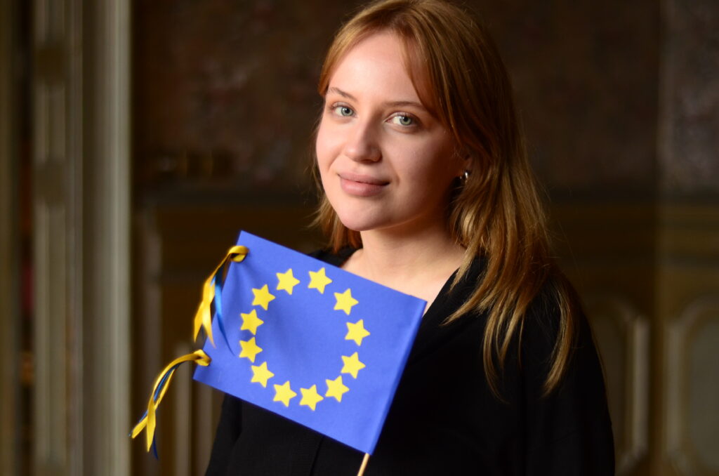 Zdjęcie przedstawia kobietę trzymającą flagę Unii Europejskiej.