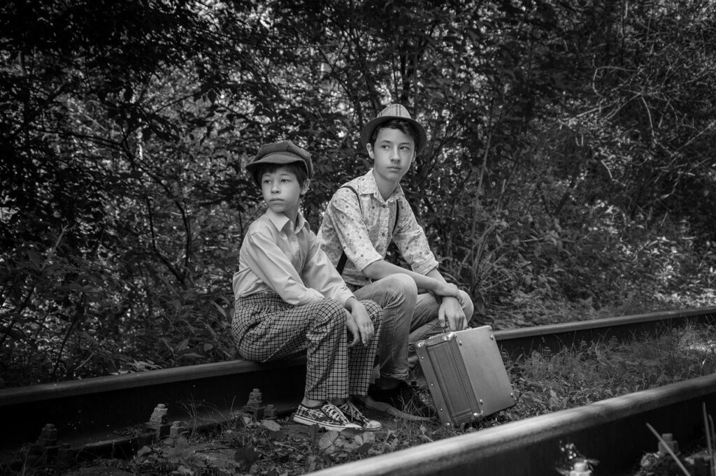 Czarno-białe zdjęcie przedstawia dwóch młodych chłopców, którzy siedzą na torach kolejowych. Jeden z nich trzyma walizkę.
